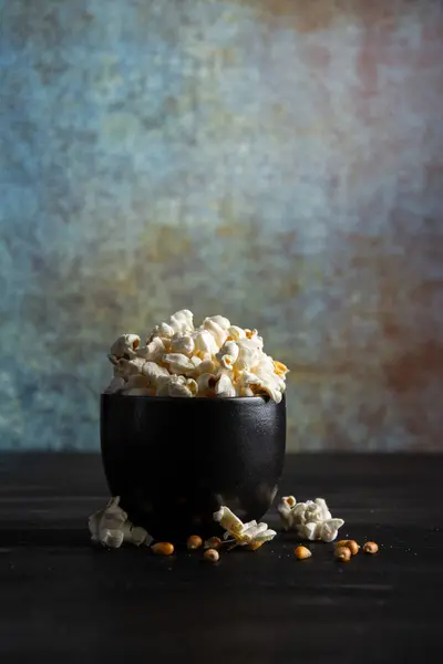 Close-up of black bowl with popcorn on dark table with popcorn, and grains, gray background, vertical, with copy space