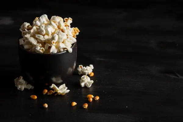 Top view of black bowl with popcorn on dark table with popcorn, grains and salt, horizontal, with copy space