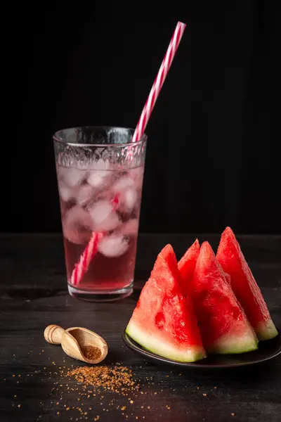 Tabağın yakın plan karpuz parçaları ile kahverengi şeker ve bir bardak meyve suyu ile buz, siyah arkaplan, dikey, kopyalama alanı ile