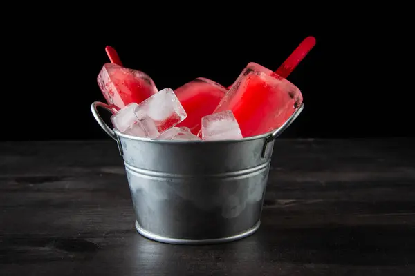 View of metal bucket with watermelon and ice lollies, on table and dark background, horizontal, with copy space