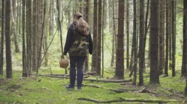 A girl in a camouflage jacket walks through the woods with a basket. Looking for mushrooms.