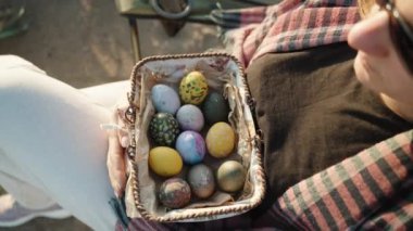 Close-up of a girl who is sitting and holding a basket with Easter colored eggs. The rays of the sun. Tilt.