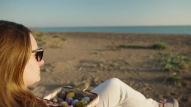 A girl is sitting on the beach at sunset with a basket of Easter Eggs in her hands and looking at the sea.