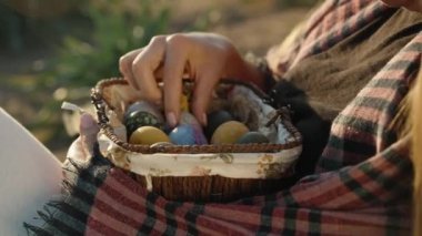 The girl is sitting in the sun holding a wicker basket with Easter eggs and twirling them with her hands. close-up.