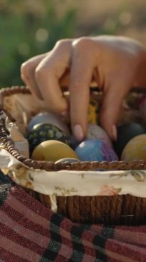 The girl is sitting in the sun holding a wicker basket with Easter eggs and twirling them with her hands. close-up. Vertical video Social Media