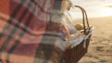 A woman carries a wicker basket with Easter Eggs, The setting sun and the wind blows the plaid on it. Sea sand and waves in the background. Slow motion.