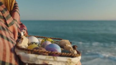 A wicker basket with Easter eggs in the hands of a girl, she is standing in the water, sea waves in the background.