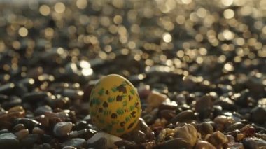 A painted Yellow Egg for Easter, on the rocks by the sea. The focus shifts to the waves and mountains behind.