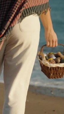 A woman with a basket of Easter Eggs walks on the sand, stops and looks into the sea. Vertical video Social Media