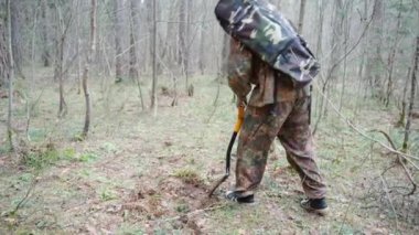 A man dressed in camouflage clothing walks through the woods with a metal detector and a shovel. Search for treasure, coins, metal.