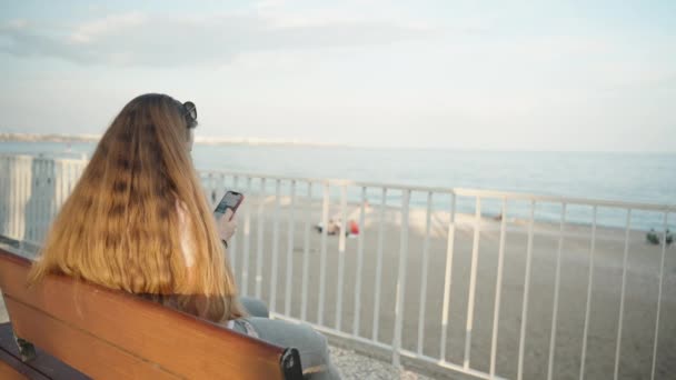 若い女性はベンチに座って 電話で写真をスクロールし 海の海の遊歩道 — ストック動画