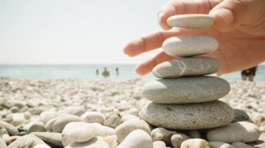 Yakın plan, bir kadın cairn yapıyor. Deniz kıyısında el yapımı taş kule. Sahilde bir yığın dengeli çakıl taşı. Denizin arka planına ve insanların yüzmesine karşı