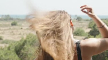 The young woman blows her long hair in the wind. She looks at the sea in the distance. Slow mo