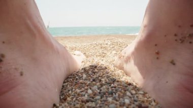 Women's feet step on small sea stones, close-up. The sea in the background.