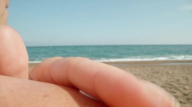 The young woman smears the burned shoulder with sunscreen against the background of the sea. Sand. Big waves. Dolly slider extreme close-up. Laowa Probe