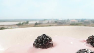Breakfast under the open sky on the balcony on a sunny morning. I toss mulberries into a fruity yogurt breakfast. Dolly slider extreme close-up. Laowa Probe. Slow mo