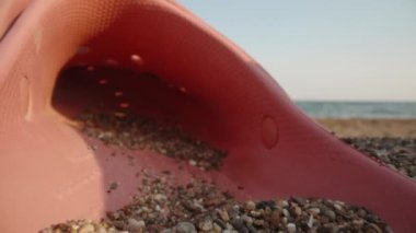 Sunglasses and pink flip flop lie in the sand against the background of the sea. The sun is reflected in glasses. Dolly slider extreme close-up. Laowa Probe.