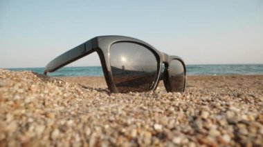 Sunglasses lie in the sand against the background of the sea. The sun is reflected in glasses. Close-up. Laowa Probe.