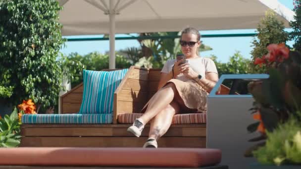 Young Woman Sits Wooden Bench Large Umbrella Looks Her Phone — Stock Video