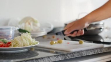 Yemek pişirirken, zeytinleri büyük bir bıçakla kesiyorum, hareket halinde. Siyah taş tezgah. Beyaz kesme tahtası. Sebzeli tabak..