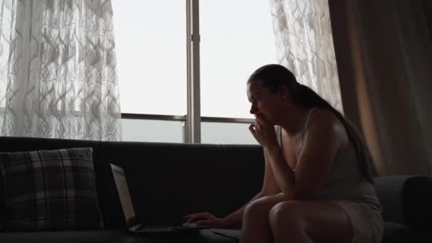 若い女性はノートパソコンで働いている 遠くの仕事だ ソファに座って 壁一面窓 動いてる — ストック動画
