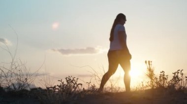 Gün batımı güneşi. Genç kadın kum ve bitkilerden oluşan bir tepede yürüyor. Güneşin arka planında