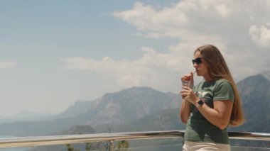 Uzun saçlı genç bir kadın buzlu kahve içer. Arkasında dağlar var. Hareket halinde.