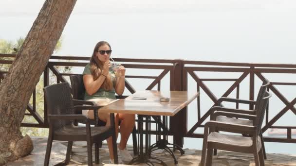 Młoda Kobieta Okularach Słonecznych Siedzi Przy Stole Cieniu Drzewa Pije — Wideo stockowe