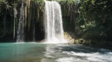 Şelale bir dağ nehrine akar. Güneş, turkuaz suyu aydınlatır. Yaz, etrafta yeşil ağaçlar. Panorama.