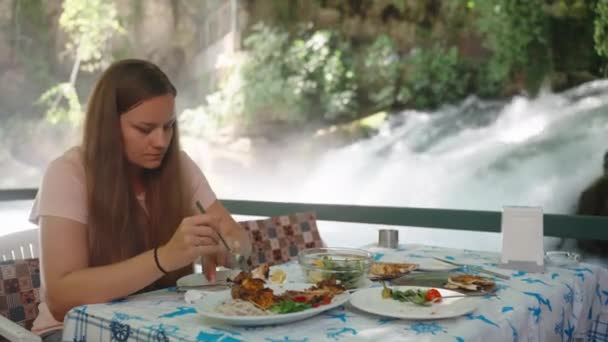 若い女性はケバブを皿に入れます 水の上のカフェで昼食 山の川の小さな滝 — ストック動画