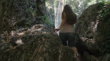 Genç kadın şelaleye doğru gidiyor. Büyük kayaların içinden geçiyor. Dikiz aynası. Yaz yürüyüşü.