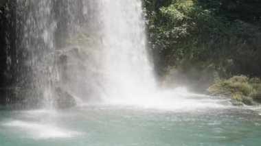 Şelale kayaya çarpıyor. Güneş, turkuaz suyu aydınlatır. Yakın plan..