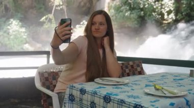 Uzun saçlı genç kadın selfie çekiyor. Telefonundaki fotoğrafa bakıyor. Arka planda bir şelale var. Suda bir kafe.