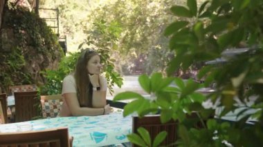 Deniz kenarındaki bir kafede genç bir kadın. Oturup başka tarafa bakıyor. Çevrede her şey yeşil. Yaz yürüyüşü.