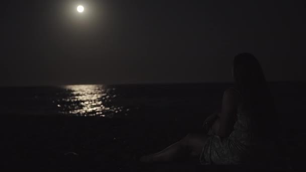 一个穿着连衣裙的年轻女子坐在海滩上的海边 她指着红色的月亮 水面上的月光 — 图库视频影像
