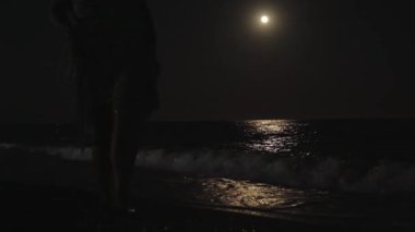 Genç kadın gece ay ışığında deniz plajı boyunca yürür. Sudaki ay ışığı, dalgalar.
