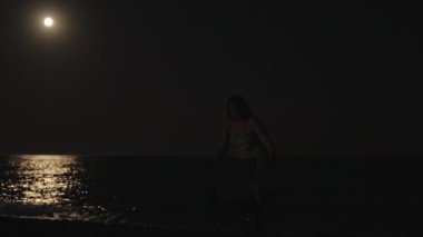 Genç kadın deniz kenarında bir sandalyede oturur ve aya bakar. Bir sandalye kapar ve gider. Ay suyu aydınlatır.
