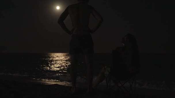 月亮和大海 那个年轻女子坐在岸上的一张椅子上 旁边站着一个男人 然后走了 — 图库视频影像