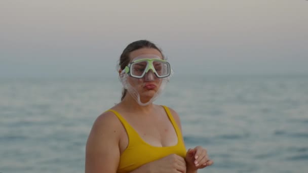 一位体形丰满的年轻女子带着面具 在夕阳西下的天空和大海的衬托下 试图用水肺潜水 做了个鬼脸 — 图库视频影像