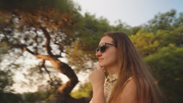 一位戴着太阳镜的年轻女子站在森林里看着夕阳西下 相机在她身边旋转着 — 图库视频影像