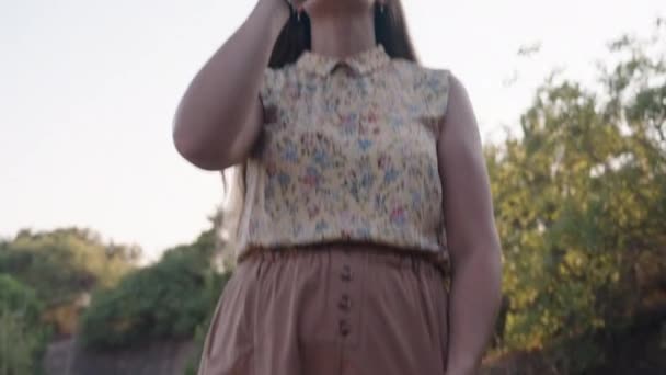 一位穿着短裙和太阳镜的年轻女子站在树后喝水 夕阳西下由下到上的移动 — 图库视频影像