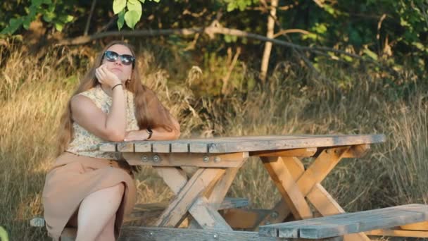 Młoda Kobieta Okularach Słonecznych Siedzi Ławce Przy Stole Tle Drzew — Wideo stockowe