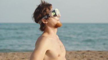 Kumsalda kızıl sakallı bir adam dalgıç gözlüğü takıyor, güneşe ve kameraya bakıyor. Arka planda deniz var. Yavaş çekim