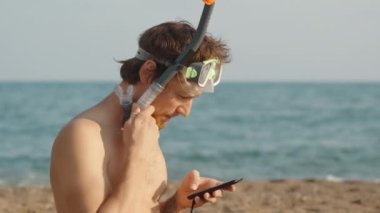 Kumsalda kızıl sakallı, dalgıç gözlüklü ve piposu olan bir adam telefona bakar ve kavun yer. Arka planda deniz var. Yavaş çekim