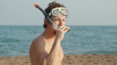 Kumsalda kızıl sakallı, dalgıç gözlüklü ve tüplü bir adam tüpte beyaz bir lolipop emiyor. Arka planda deniz var. Yavaş çekim