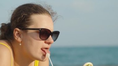 Tombul genç bir kadın kumsalda uzanıyor ve sırayla beyaz ve renkli bir lolipopu yalıyor. Sarı bir mayo ve bardakla, arka planda deniz. Yaklaş, ağır çekim.