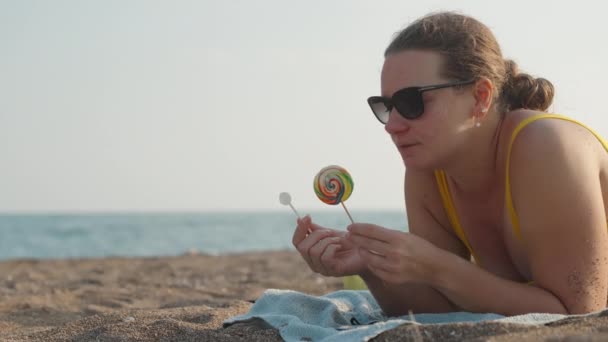 Chubby Young Woman Lies Beach Chooses Two Lollipops Yellow Swimsuit — Stock Video