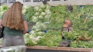 Sebze dükkanındaki genç bir kadın farklı sebze ve salata seçer. Aynalı vitrinler.