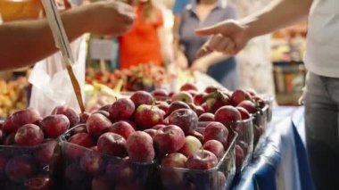 Bir sebze pazarı, kırmızı elmalı bir dükkan, satıcı alıcıdan bir elma alır ve bir torbaya koyar..