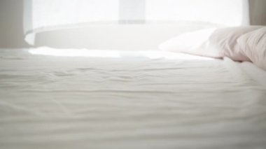 Güneşli bir sabah, pencereden gelen ışık beyaz yatağa düşer, rüzgar perdeleri savurur. Yataktan pencereye genel bakış.